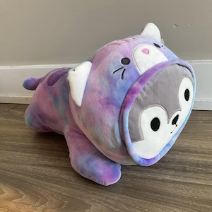 May include: A plush toy in the shape of a seal, featuring a tie-dye pattern in shades of purple, blue, and pink. The seal wears a hooded costume with a gray face and black eyes. The toy has white accents and is lying on a wooden surface.