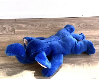 May include: A plush, cobalt blue elephant toy lying on a wooden surface. The elephant has large ears with pink inner ears and a soft, furry texture. The toy is in a relaxed pose.