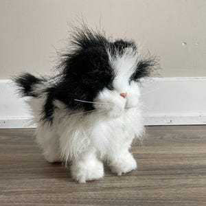 Op de afbeelding: Een pluche kat met zwart en wit haar. De kat heeft een pluizig uiterlijk met een zwarte vlek op zijn hoofd en rug, en witte vacht op zijn gezicht, borst en poten. Het heeft een roze neus en lange witte snorharen.