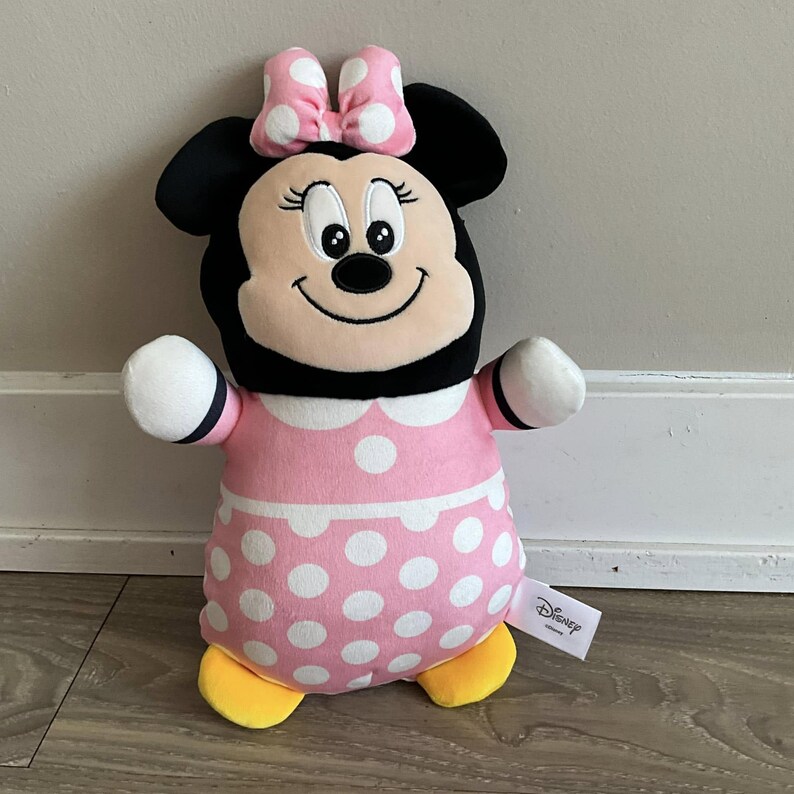 K&ouml;nnte beinhalten: Ein rosa und schwarzes Pl&uuml;schtier von Minnie Maus mit einem wei&szlig;en, gepunkteten Kleid und gelben Schuhen. Das Spielzeug hat eine schwarze Schleife mit wei&szlig;en Punkten auf dem Kopf. Das Spielzeug hat ein Disney-Logo-Etikett.