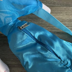 May include: A close-up of a light blue satiny bag with a zipper closure and a blue strap. The bag has a silver zipper and a white interior. The bag is likely a children's accessory.