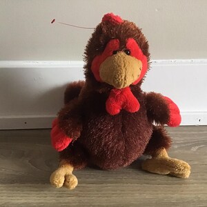 Puede incluir: Un juguete de peluche de pollo marrón y rojo con una cresta y una papada rojas. El pollo tiene un cuerpo marrón con acentos rojos en sus alas y pies.