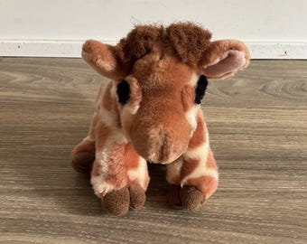 Könnte beinhalten: Ein Plüsch-Giraffen-Spielzeug mit einem braunen und cremefarbenen Fleckenmuster. Die Giraffe hat braune Hufe, schwarze Augen und einen Büschel braunes Fell auf dem Kopf. Das Spielzeug liegt auf einer Holzoberfläche.