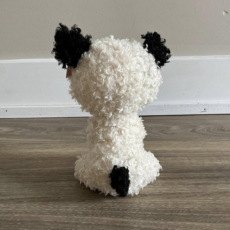 May include: White plush panda bear with black ears and a black tail. The panda is sitting on a wooden floor.