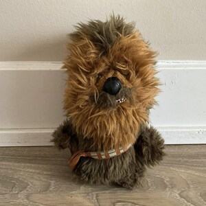 Op de afbeelding: Een pluche speelgoed dat een personage afbeeldt met bruine en bruine vacht, een zwarte neus en een bandolier. Het speelgoed heeft een gedetailleerd gezicht met kleine tanden en staat rechtop. Het speelgoed is een Star Wars-personage.