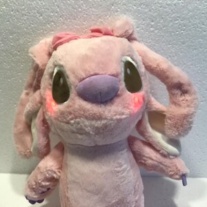 May include: Pink plush toy with a purple nose and brown eyes. The toy has a pink bow on its head and is wearing a white collar.