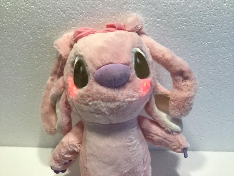 May include: Pink plush toy with a purple nose and brown eyes. The toy has a pink bow on its head and is wearing a white collar.