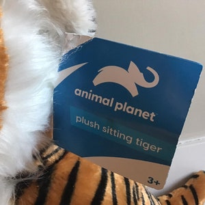 May include: A plush sitting tiger toy with orange and black stripes and white fur. The tag reads "animal planet plush sitting tiger 3+"