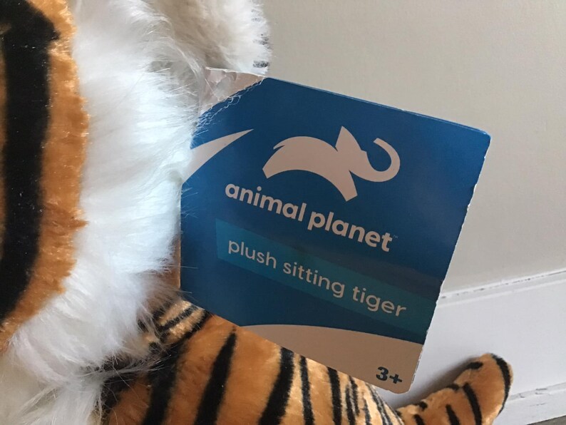 May include: A plush sitting tiger toy with orange and black stripes and white fur. The tag reads "animal planet plush sitting tiger 3+"