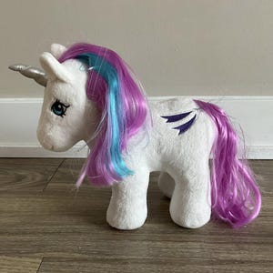 May include: A white plush unicorn toy with a silver horn and blue eyes. The unicorn has a purple and blue mane and tail, and purple wing accents on its side. The toy is made of soft material.