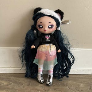May include: A Na! Na! Na! Surprise doll with long dark blue hair and a panda bear hat. The doll is dressed in a black hoodie, a colourful skirt, and white boots with black accents. The doll has large, expressive eyes and is posed against a neutral wall.