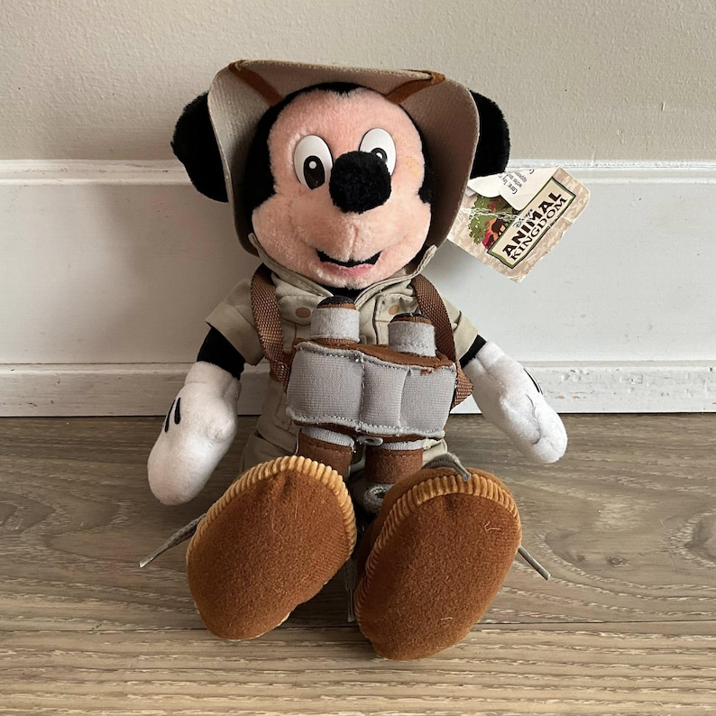 May include: A plush Mickey Mouse toy dressed in a tan safari outfit, including a matching hat and a vest. The character wears brown boots and has a pair of grey binoculars hanging around the neck. A tag labeled Disney Animal Kingdom is attached.