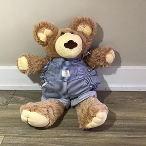 Op de afbeelding: Een bruine teddybeer met een blauw-witte gestreepte overall. De beer heeft een lichtbruine snuit en poten, met een kleine patch op de overall. De beer zit op een houten vloer.
