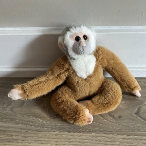 May include: A plush monkey toy with brown fur, a white face, and a gray head. The toy is sitting with its arms outstretched and its legs crossed. The monkey has dark eyes and pink hands and feet.