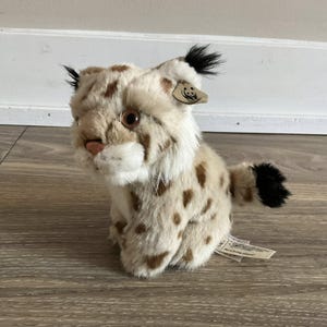 May include: A plush lynx toy with a cream and brown spotted pattern. The stuffed animal has black tufts on its ears and a black-tipped tail. It has a pink nose and brown eyes. A tag is attached to the ear.
