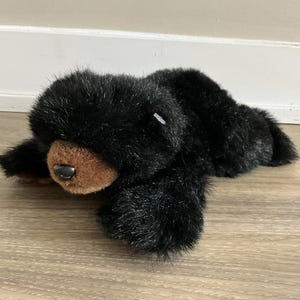 May include: A black plush bear with a brown nose and eyes. The bear is lying on its side with its head resting on its paws.