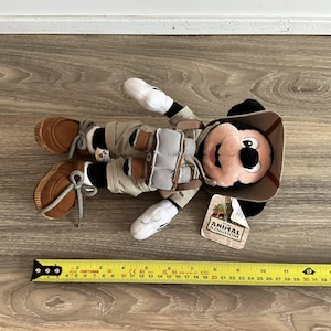 May include: A plush Mickey Mouse toy dressed in a safari outfit with a tan hat, khaki shorts, and brown shoes. The toy wears a harness with grey binoculars and has a tag reading Disney Animal Kingdom. A yellow ruler shows it is 11 inches long.