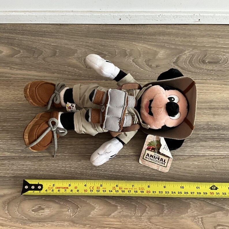 May include: A plush Mickey Mouse toy dressed in a safari outfit with a tan hat, khaki shorts, and brown shoes. The toy wears a harness with grey binoculars and has a tag reading Disney Animal Kingdom. A yellow ruler shows it is 11 inches long.