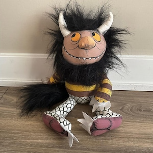 May include: A plush monster doll featuring a round tan face with yellow eyes, a wide grin, and two white horns. It has a thick black faux fur mane, a brown and yellow striped torso, and white legs with a black scale pattern and pinkish-brown feet.