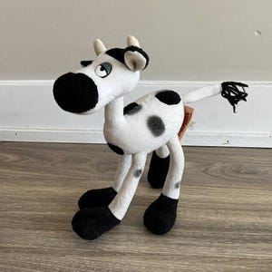 May include: A soft toy cow in a black and white spotted design. The toy features a large black snout, black hooves, and a black tail. The cow has small horns and a tag attached to its side.