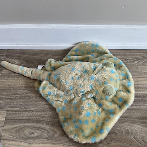 May include: A soft toy stingray in tan and blue. The toy has a rounded body, a long tail, and is covered in blue spots. The plush toy has a cuddly appearance, suitable for children.