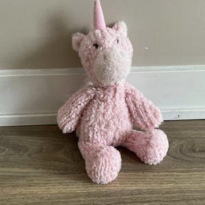 May include: A plush pink unicorn toy with a soft, textured body, a white muzzle, and a pink horn. The stuffed animal is sitting on a wooden floor, leaning against a white wall.