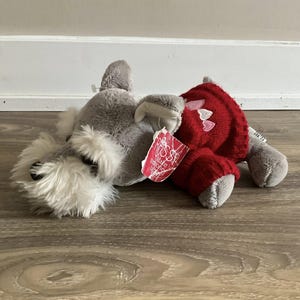 Könnte beinhalten: Ein graues Plüschhundespielzeug mit einem roten Pullover, der mit rosa Herzen verziert ist. Der Hund hat einen flauschigen weißen Bart und ein Etikett mit der Aufschrift "Make Someone Happy". Das Spielzeug liegt auf einer Holzoberfläche.