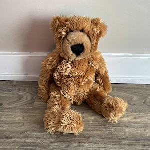 Könnte beinhalten: Ein brauner Teddybär mit schwarzer Nase und weichem, flauschigem Fell. Der Bär sitzt auf einem Holzboden und lehnt an einer weißen Wand. Das Fell des Bären hat eine warme, goldbraune Farbe.