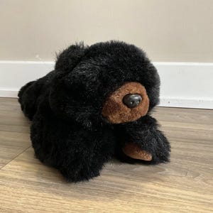 May include: A black plush bear toy with a brown snout and paws. The bear is lying down on a wooden floor. The bear's fur is soft and fluffy, and the toy has a realistic appearance.