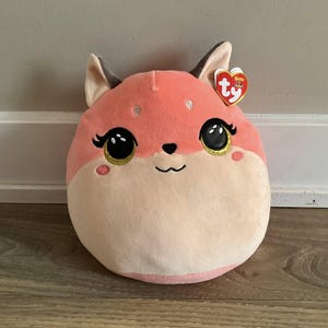 May include: A soft, plush fox toy in a rounded shape. The toy has a pink and cream colour scheme, with large black eyes outlined in gold, and a small black nose. A red heart-shaped tag is attached to the ear.