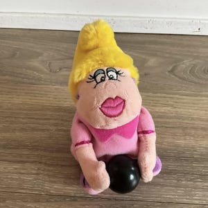 Può includere: Una bambola di peluche rosa e gialla, che tiene una palla nera. La bambola ha un cappello giallo, pelle rosa e grandi labbra rosa. Indossa una camicia rosa con un fiocco e accenti viola.