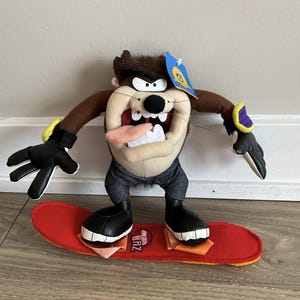 May include: A plush Taz figure from Looney Tunes, standing on a red skateboard. The figure has a tan face, brown hair, and black gloves. The skateboard has the letters "TAZ" printed on it.