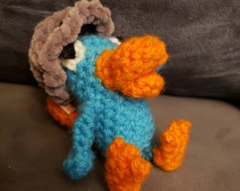 May include: A blue and orange crocheted plush toy resembling a duck wearing a gray crocheted hat. The toy is sitting on a gray surface.