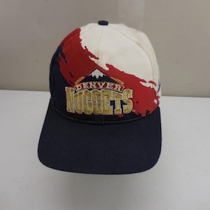 Könnte beinhalten: Eine Baseballkappe mit marineblauem Schirm und Seitenteilen sowie einem weißen und roten Pinselstrich-Design auf der Krone. Die Vorderseite zeigt ein gesticktes Denver Nuggets-Logo in Gold, Weiß und Marineblau mit einer Berggipfel-Grafik.