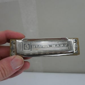 May include: A silver-colored metal harmonica with a tan wooden comb. The top cover plate features the text "BLUES HARP" and the numbers 1 through 10 engraved along the bottom edge. A small square logo is visible on the left side of the plate.