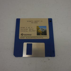May include: A blue 3.5-inch floppy disk for the game King's Quest II MS-DOS. The label is tan with black text, featuring the Sierra logo, version numbers, and a small square image of a mountain landscape. The serial number 3126289062703976 is printed.
