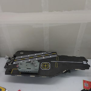Pode incluir: Um porta-aviões de brinquedo de plástico preto com uma torre de comando cinza, marcações de pista brancas e amarelas e o número 60 impresso em branco no convés. O convés de voo inclui um gráfico de zona de aterragem com uma cruz amarela.