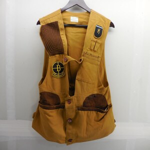 May include: A mustard yellow shooting vest featuring a brown quilted shoulder patch, a black and yellow Bushnell patch, a Browning logo patch, and the embroidered name John Heinrich. The vest includes multiple front pockets and button closures.