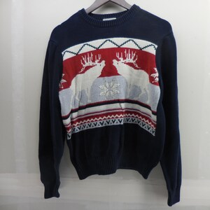 May include: A navy blue long-sleeve crew neck jumper featuring a central knit pattern of two white deer facing each other, set against a red and light blue background with a central snowflake motif and geometric border designs at the hem.