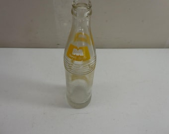 Könnte beinhalten: Eine klare Glasflasche mit engem Hals und abgerundetem Körper. Die Flasche hat ein gelbes Etikett mit weißem Text, auf dem Root Beer steht. Um den oberen Teil des Flaschenkörpers sind horizontale Rillen in das Glas eingeprägt.