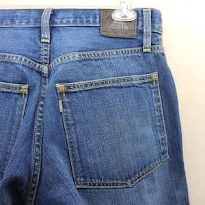 May include: Close-up of the back of blue denim jeans. The jeans have a classic five-pocket design with yellow stitching. A leather patch with the brand name "SILVERTAB" is visible at the waistband.
