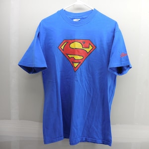 Könnte beinhalten: Ein leuchtend blaues Kurzarm-T-Shirt mit dem ikonischen roten und gelben Superman-S-Schild-Logo mittig auf der Brust. Das T-Shirt hat einen Rundhalsausschnitt und ein kleines rotes Schriftlogo auf dem linken Ärmel, präsentiert auf einem weißen Bügel.