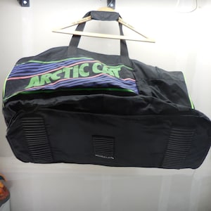 May include: A black duffel bag featuring the text ARCTIC CAT in large green letters. The bag has horizontal purple and blue stripes above the text, black carrying handles, and three black rectangular textured pads on the bottom exterior surface.