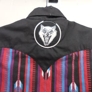 May include: A black button-down shirt featuring a pointed western-style yoke and a circular patch on the back. The patch displays a white graphic of a snarling wolf head. The lower portion of the shirt has a vertical striped pattern in red, blue, and purple.