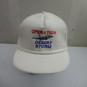 May include: A white corduroy baseball cap featuring an embroidered graphic of a fighter jet. Above the jet, the word OPERATION is stitched in red, and below the jet, the words DESERT STORM are stitched in blue. A white braided cord trim sits above the brim.