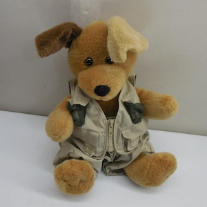 Puede incluir: Un perro de peluche con pelaje marrón, que presenta una oreja de color marrón oscuro y otra de color beige claro. El animal de peluche lleva un chaleco utilitario beige con cremallera frontal, múltiples bolsillos y detalles de malla en el pecho.