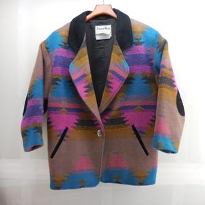 May include: A long-sleeve blazer featuring a vibrant geometric pattern in shades of blue, magenta, and tan. The garment includes a solid black collar, black elbow patches, two front pockets with black trim, and a single button closure. A Pioneer Wear label is visible.