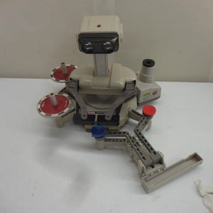 May include: A beige robotic toy with a head featuring two black lenses, a central body with gray articulated arms, and two red circular trays. The base includes a long gray extension arm with a red button and a blue button, plus a small tray.