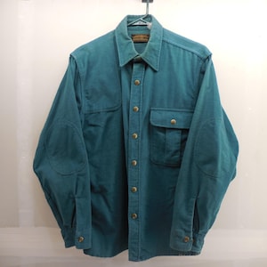 May include: A teal long-sleeve button-down shirt featuring a pointed collar, a single chest pocket with a flap, and reinforced elbow patches. The shirt includes metallic buttons down the front placket and on the cuffs, with a visible brand label.