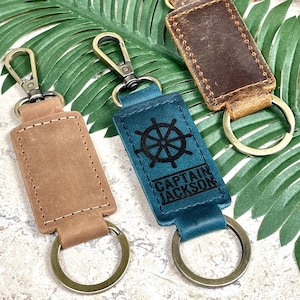 May include: Three leather keyrings with metal rings. One keyring is brown with a blank space for personalisation. One keyring is blue with a ship's wheel design and the text "Captain Jackson". One keyring is brown with a distressed leather look.